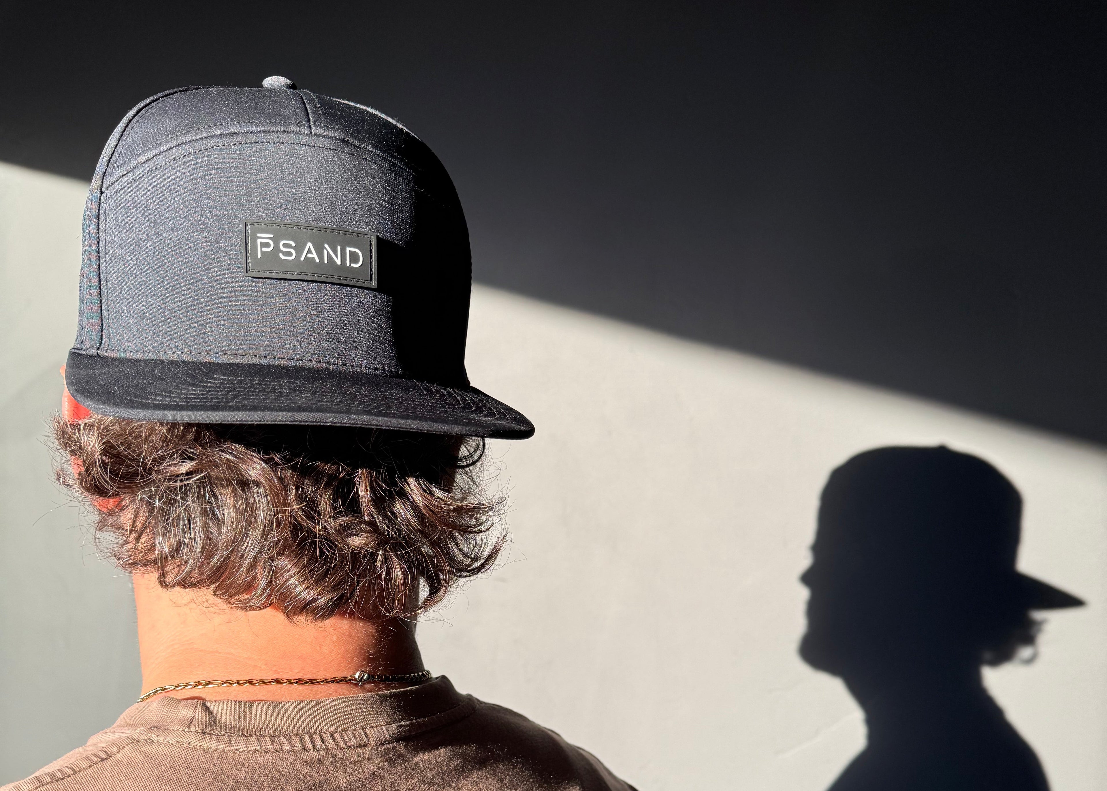 Person wearing a black hat with 'PSAND' logo, casting a shadow on a light background